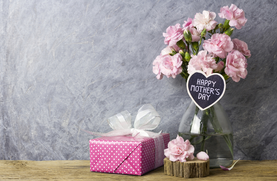 shutterstock_544309597 Blumenstrauß mit rosa Nelken in einer Glasvase, beschriftet mit "HAPPY MOTHER'S DAY", neben einem pinken Geschenk mit weißen Punkten auf einem Holztisch, passend zum Muttertag und zu RAU Cosmetics' Angeboten für Pflegeprodukte.