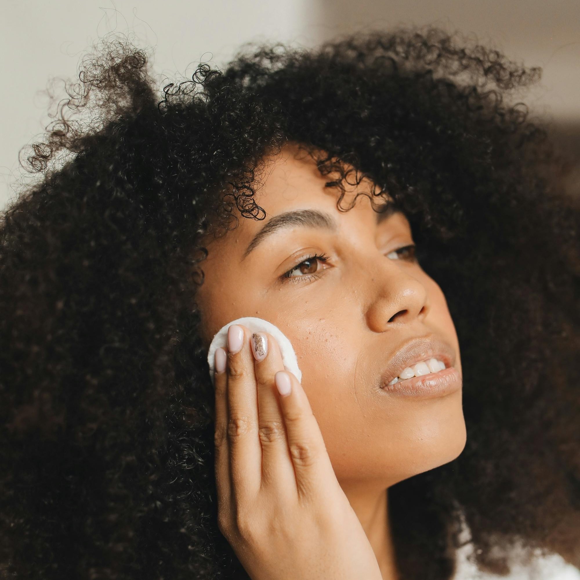 Frau mit lockigem Haar reinigt ihr Gesicht mit einem Wattepad, symbolisiert die Hautpflege und die Vorteile des Hydro Facials von RAU Cosmetics.