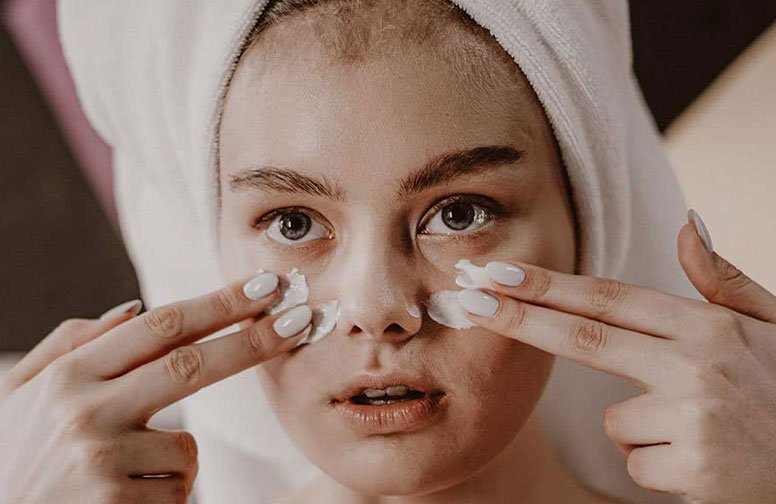 Frau mit Handtuch im Haar, die Gesichtscreme auf die Wangen aufträgt, symbolisiert die Anwendung von Hautpflegeprodukten und das Thema Pilling-Effekt in der Gesichtspflege.