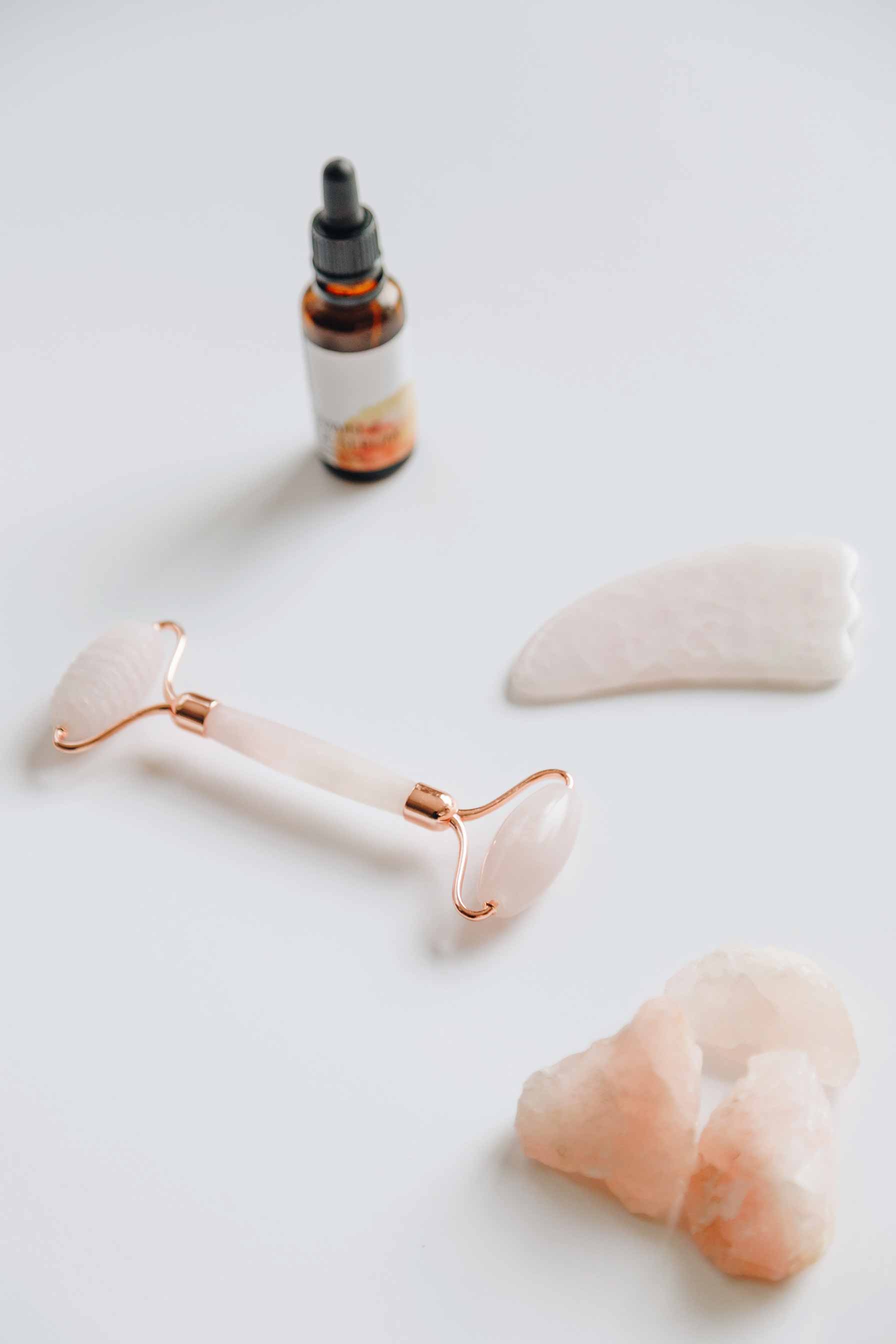 hautpflege-trends-2020 Rosenquarz Gesichtsroller, Gua Sha Stein und Hautpflegeöl auf weißem Hintergrund, symbolisierend die Hautpflege-Trends 2020 und natürliche Beauty-Rituale.