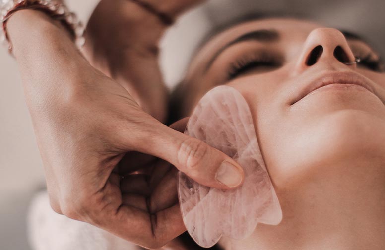 rau-cosmetics-gua-sha-massage-02 Gua Sha Gesichtsmassage mit Rosenquarzstein, Hand führt sanfte Massagebewegungen über das Gesicht einer Frau, Hautpflege und Entspannung.