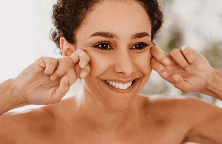 Haut-Gesicht-Wassereinlagerungen-02 Frau mit fröhlichem Gesichtsausdruck, die ihre Wangen mit den Händen zieht, zur Veranschaulichung von manueller Lymphdrainage zur Reduzierung von Wassereinlagerungen im Gesicht.
