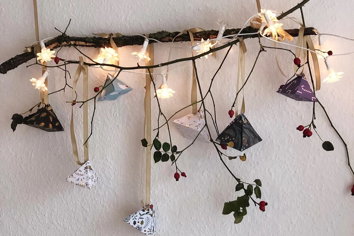 Selbstgemachter Adventskalender aus dekorativen Pyramiden, aufgehängt an einem Ast mit Lichtern und Zweigen, ideal für kreative Weihnachtsüberraschungen.